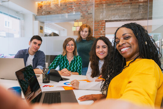 A multiracial group of young professionals smiles at the camera while taking a selfie in a modern office. Friendly and collaborative work environment