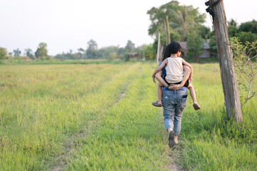 Adult carrying a young boy on their back while walking through a green rural field under the open sky.