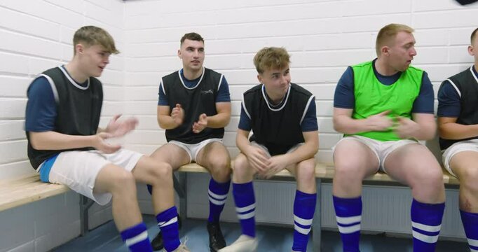 Male soccer players wearing black vests and tying shoes sitting on bench in changing room with coach