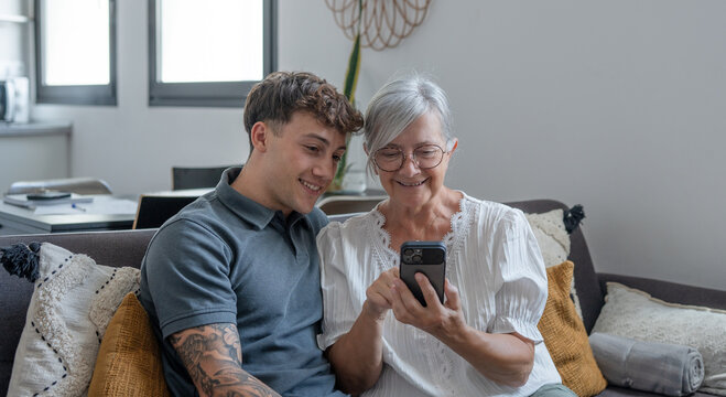 Happy grandmother learning how to use mobile phone technology from young grandson, handsome smiling boy teaching grandma shop online, book travel and vacation