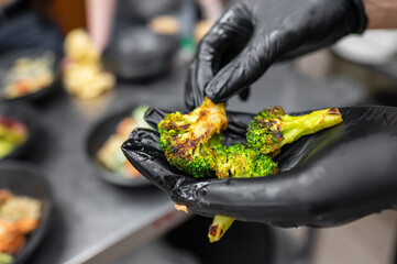A chef in black gloves holds a piece of roasted broccoli, showcasing its vibrant green color and charred edges, against a backdrop of prepared dishes on a kitchen countertop.
