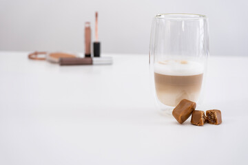 A close up shot of a stylish glass of layered coffee with a creamy foam top, accompanied by caramel chocolate candies, one of which is broken in half revealing a rich filling. 