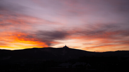 A winter evening casts vibrant colors across the sky above Jested Mountain in Czechia. The silhouette of the mountain stands against a stunning backdrop of orange and purple hues.