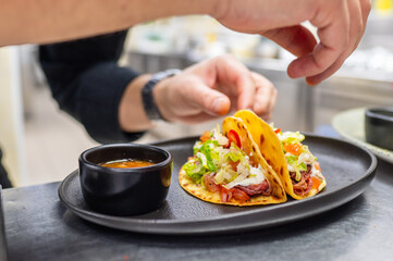 A chef garnishes two delicious tacos filled with fresh ingredients, served on a sleek black plate alongside a small bowl of sauce. Perfect for culinary or food-themed projects.