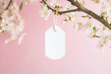 Blank white paper tag mockup hanging on blooming spring branch on pastel pink background