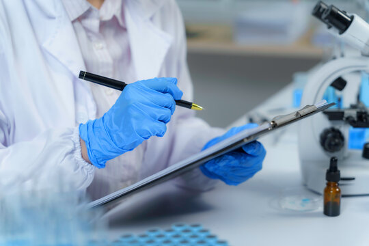 Scientist wearing lab coat and gloves taking notes on clipboard in laboratory setting
