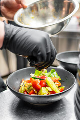 A close-up of a chef in black gloves preparing a fresh salad, adding dressing to a vibrant mix of lettuce, tomatoes, and cucumbers in a stylish bowl in a professional kitchen setting.