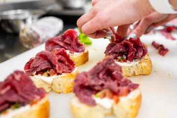 A close-up of a chef's hands adding slices of cured meat onto toasted bread with creamy spread and fresh toppings, showcasing culinary preparation in a professional kitchen.