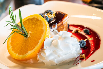 A moist blueberry-topped muffin paired with a vibrant orange slice, garnished with rosemary, and complemented by a drizzle of berry sauce for a refreshing finish.
