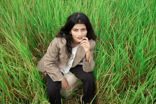 a young Indian female model with black hair, wheatish skin, Indian features wearing a brown coat over a white t shirt and black trousers posing middle of an open grass field