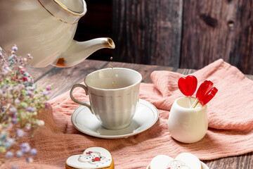 a woman pours tea from a white teapot into a white porcelain cup with a saucer, next to gingerbread cookies with white glaze, red heart-shaped lollipops, Valentine's Day composition, Valentine's Day t