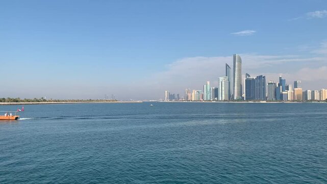 Panoramic, scenic view of Abu Dhabi city skyline and waterfront. Traditional wooden abra boat is sailing in front. UAE flag is waving on boat. Al Lulu Island. Abu Dhabi, United Arab Emirates