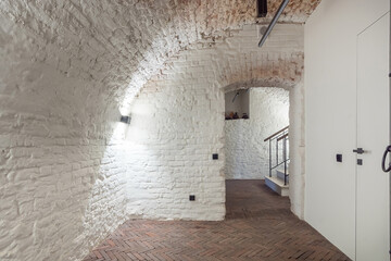 A bright, spacious hallway features white brick walls and a herringbone wood floor. Light fixtures illuminate the area, with a staircase visible beyond.
