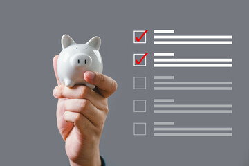 Hand holding a white piggy bank against a checklist background, symbolizing financial planning,...