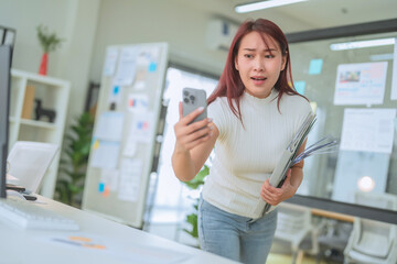 Asian woman in office looks surprised while holding phone and documents. She appears to be reacting to unexpected news or information