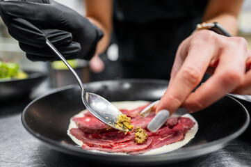 A close-up of a chef's hand adding garnishes to a plate of sliced meat. The preparation features elegant presentation, showcasing culinary artistry in a modern kitchen setting.