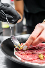 A close-up of a chef's hands in black gloves carefully applying mustard on thinly sliced beef in a modern kitchen setting. The focus is on the culinary artistry and attention to detail.