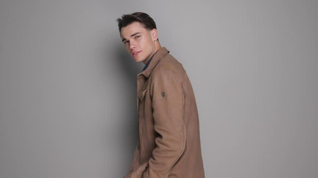 handsome casual boy rotating and fixing brown leather jacket collar while standing on gray studio background