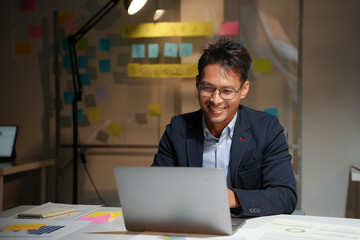 Happy entrepreneur smiling and working at night using laptop in office with sticky notes on glass board