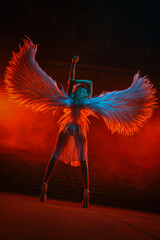 Blonde girl in a white light dress with angel wings posing on fiery smoke background.