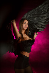 Beautiful girl in a black corset and black tights with black angel wings on pink smoky background.
