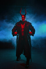 Huge muscle man with devil horns in a black coat in blue smoke on dark black background.