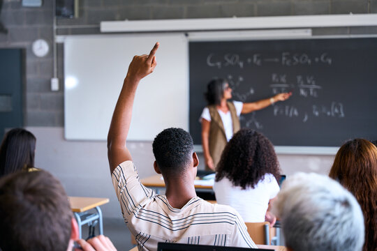 Young group of gen z students attending class at high school. Young black student boy hand raised asking a question to senior female teacher during lecture. Education concept