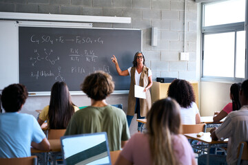 View from behind a group of multiracial students taking notes in a classroom. At the front, the teacher stands at the blackboard, giving a lesson, emphasizing focus and the exchange of knowledge
