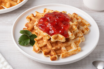 Delicious waffles topped with strawberry jam and a fresh leaf on a white plate with a light wooden background