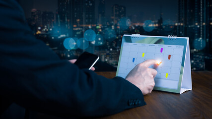 Timetable, agenda, and schedule for the 2025 event planner. A businessman at an office desk takes notes on the calendar while using his phone to check the planner. Using calendars and event organizing