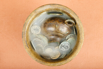 Ancient bronze pot filled with old coins and mist effect on an orange background.