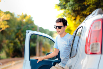 Casual traveler exiting a car during a scenic road trip. The man is dressed in light clothes, wearing sunglasses, with a natural and sunny outdoor background.