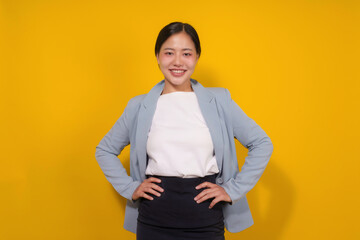 Portrait of a smiling young Asian businesswoman standing with hands on hips against a vibrant yellow background, exuding confidence and professionalism