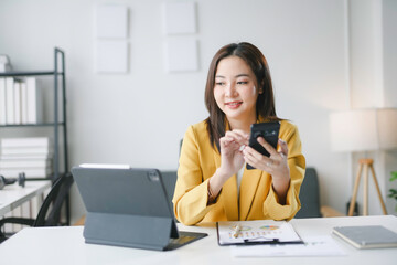 Young professional woman using smartphone and working with digital tablet and financial chart at...