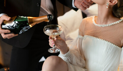 An elegantly dressed man pours champagne into a glass held by a woman in a white dress in a luxurious setting