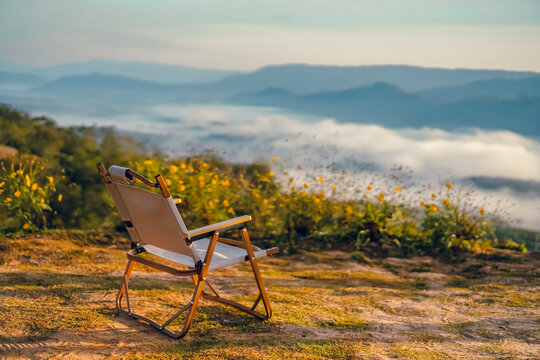 A white camping chair on the ground with a misty mountain view on a cold day in the morning. There are yellow flower bushes on green grass in soft tone. Idia for outdoor background with copy space.