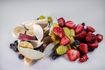 A colorful assortment of freeze-dried apples, bananas, strawberries, and blueberries displayed on a white surface. The vibrant textures and natural colors emphasize their preserved freshness.