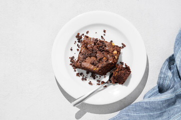 Chocolate brownie with nuts on a plate with spoon on a light background with morning shadow