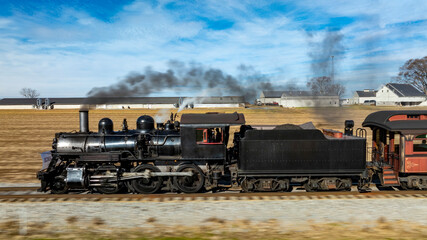 A vintage steam locomotive powers along the tracks, sending plumes of smoke into the blue sky. Surrounding fields and farmhouses enhance the picturesque countryside setting.