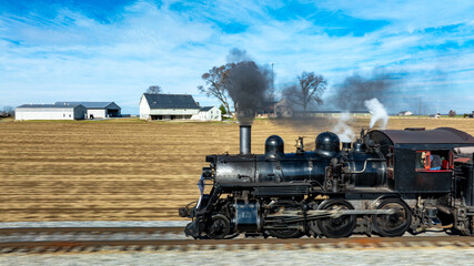 A vintage steam locomotive powers along the tracks, sending plumes of smoke into the blue sky. Surrounding fields and farmhouses enhance the picturesque countryside setting.