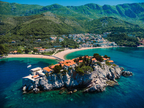 An aerial view of a coastal town with a historic fortress on rocky cliffs surrounded by turquoise waters. Sandy beaches, a yacht, and green mountains create a perfect Mediterranean landscape