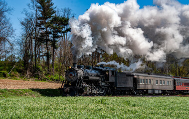 A large steam locomotive steams through a green field under blue skies, surrounded by trees. The train emits thick white smoke, creating a striking contrast with the vibrant scenery.