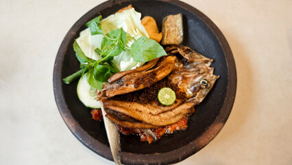 Crispy fried milkfish served with spicy sambal penyet and fresh vegetables on a rustic black plate in Indonesia featuring vibrant garnishes and traditional presentation.