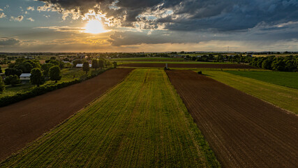 As the sun sets in a vibrant sky, vast agricultural fields stretch across the landscape, dotted with trees and farm structures, creating a serene and picturesque rural scene.
