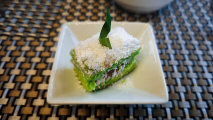 Indonesian Traditional Green Lumpang Cake with Grated Coconut Garnish Served on a White Plate Against a Textured Woven Background