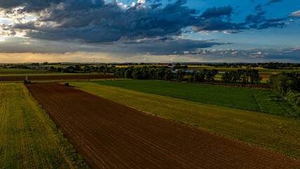 Obraz premium As the sun sets in a vibrant sky, vast agricultural fields stretch across the landscape, dotted with trees and farm structures, creating a serene and picturesque rural scene.