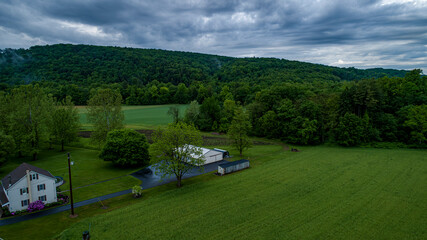 Expansive farmland showcases vibrant greenery and a distant forest against a backdrop of moody gray clouds. A small house and storage buildings add to the serene rural setting.