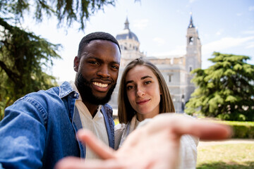 Happy multiethnic couple taking a selfie with outstretched hand in a park with the cathedral in the background