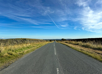 A long, empty Green Sykes road stretches towards the horizon under a blue sky, with grassy fields and stone walls lining either side. The sky is adorned with wispy clouds near, Oakworth, UK