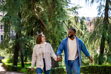 Fototapeta premium Young multiethnic couple holding hands and walking together in a green park, enjoying a romantic stroll on a sunny day